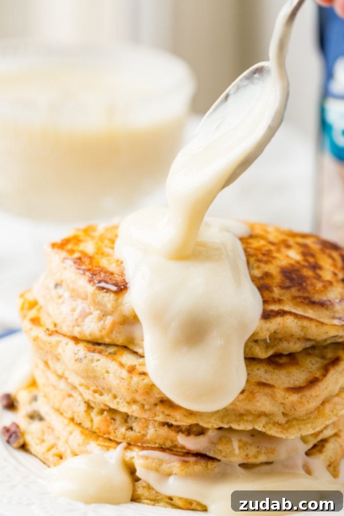Sweet and Spiced Carrot Cake Pancakes 7 A photo thick white cream cheese frosting sauce being poured onto a stack of golden carrot cake pancakes with a spoon.