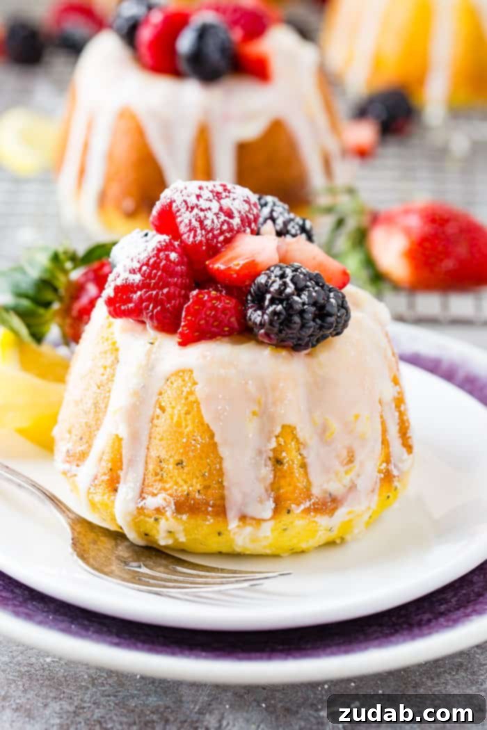 Zesty Lemon Poppy Seed Bundtlets 2 A photo of a mini lemon poppy seed bundt cake on white plate with a lemon glaze on top and fresh powder sugar dusted berries.