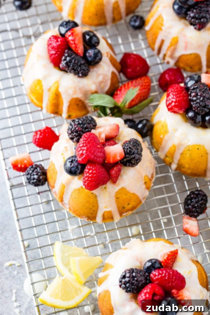 Zesty Lemon Poppy Seed Bundtlets 3 A photo of several mini lemon poppy seed bundt cakes on a silver wired cooling rack topped with lemon glaze and fresh berries.
