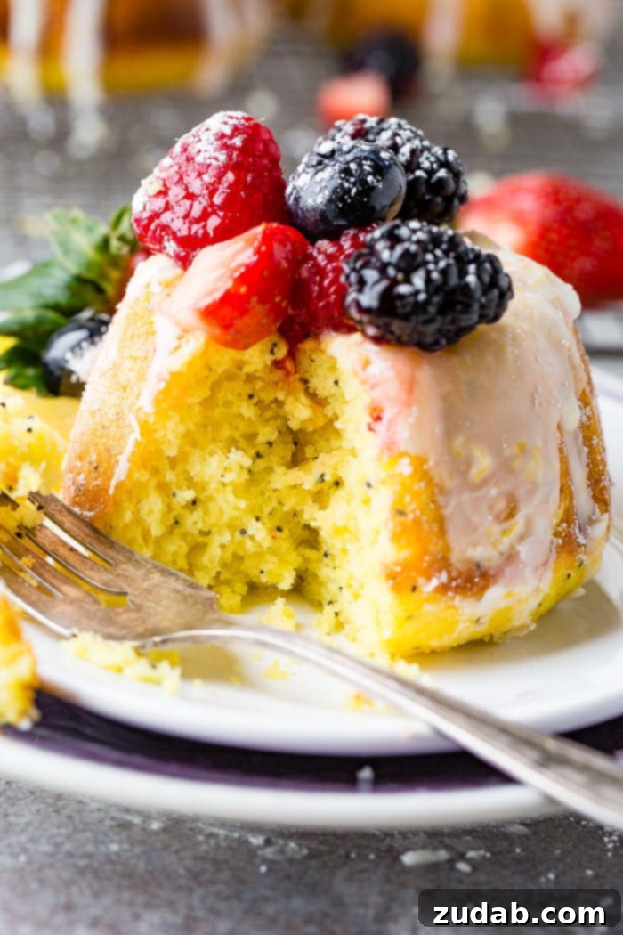 Zesty Lemon Poppy Seed Bundtlets 4 A photo of a mini lemon poppy seed bundt cake with a bite taken out on white plate with a lemon glaze on top and fresh powder sugar dusted berries.
