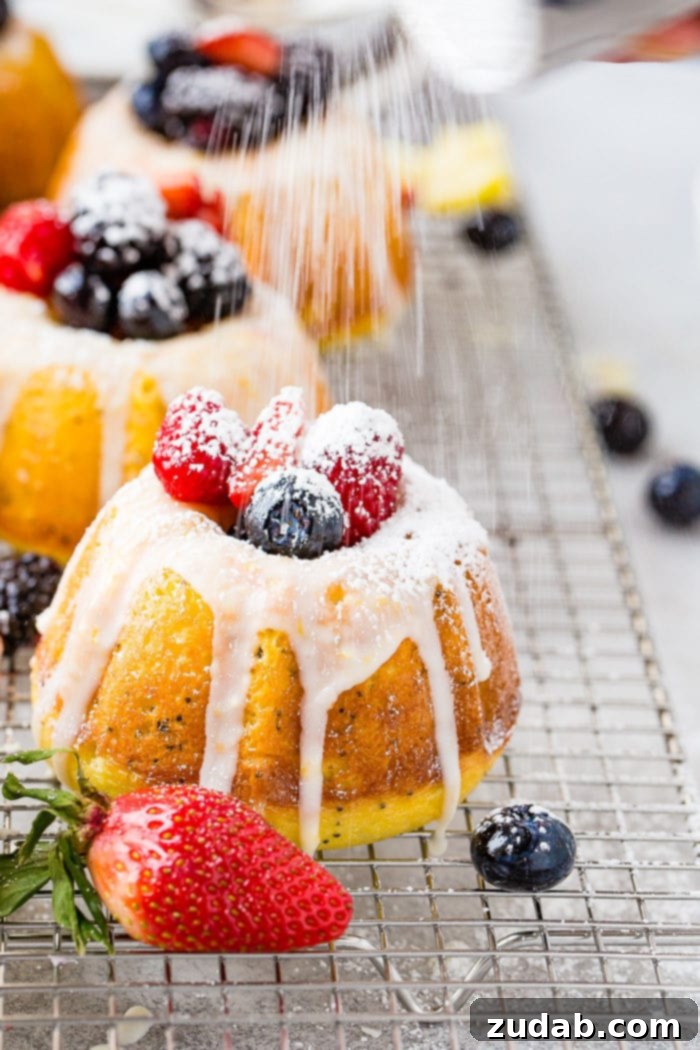 Zesty Lemon Poppy Seed Bundtlets 6 A photo of a mini lemon poppy seed bundt cake on a silver wired cooling rack with a lemon glaze on top and fresh powder sugar being dusted on fresh berries on top.