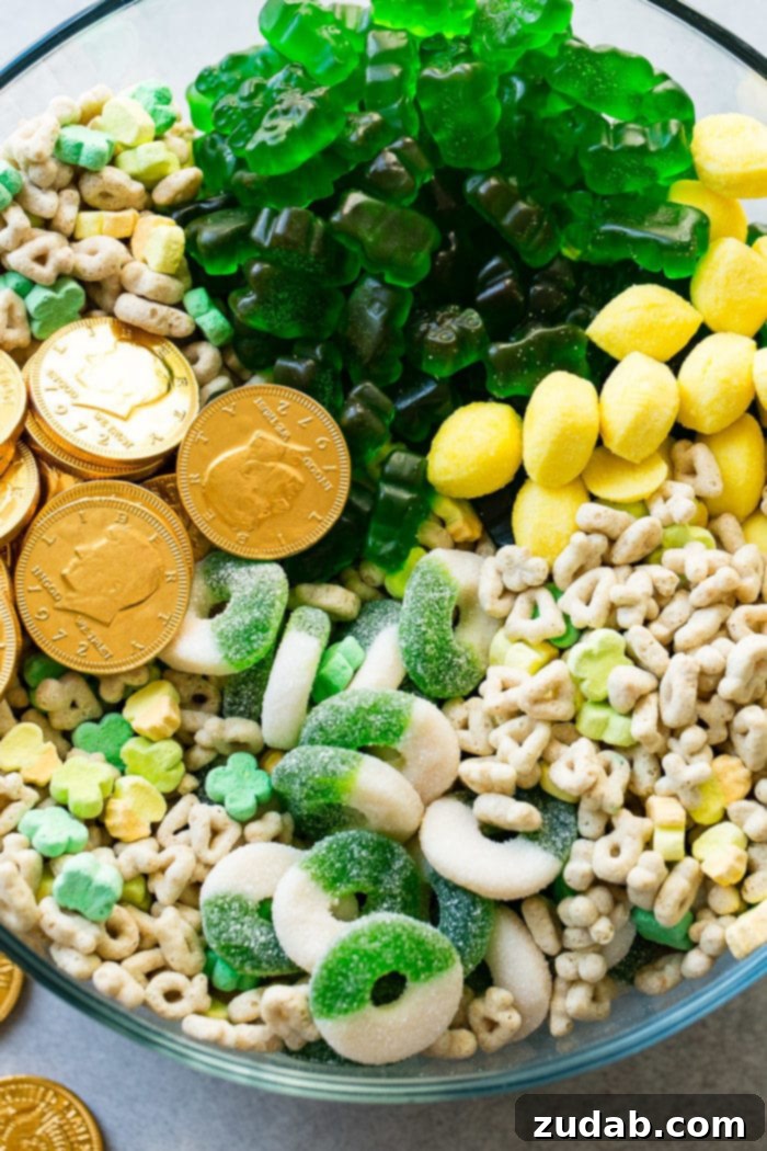 Lucky Leprechaun's 5-Minute Treat 5 A close-up photograph highlighting the tempting variety within the St. Patrick's Day snack mix in a glass bowl. Detailed view of chocolate gold coins, Lucky Charms marshmallows, bright lemon drops, and an assortment of green gummy candies, emphasizing the rich textures and colors.