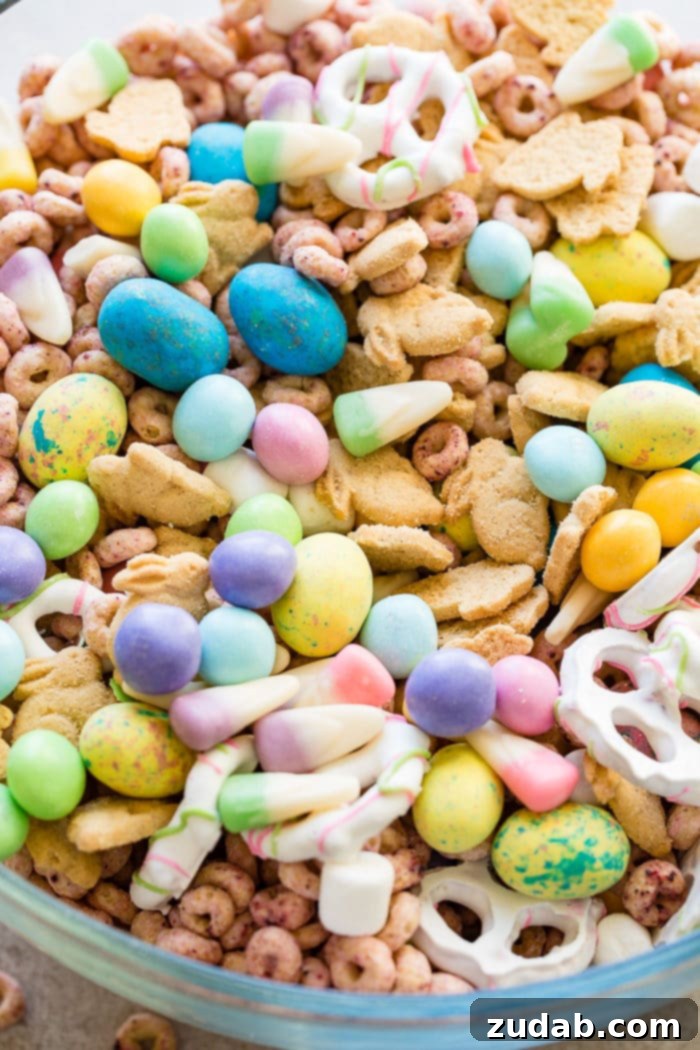 Effortless Easter Snack Medley 3 A close-up of a festive Easter snack mix showcasing a delightful blend of colorful candies, crunchy berry Cheerios, sweet mini marshmallows, and charming bunny-shaped cookies in a clear glass serving bowl.