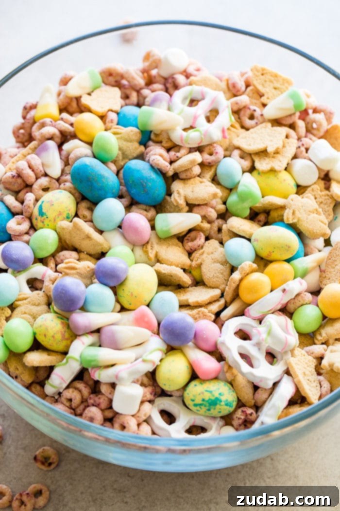 Effortless Easter Snack Medley 5 A generous clear glass bowl filled with a delightful Easter snack mix, featuring a colorful array of cereals, candies, chocolate eggs, pretzels, and bunny cookies, set against a festive background.