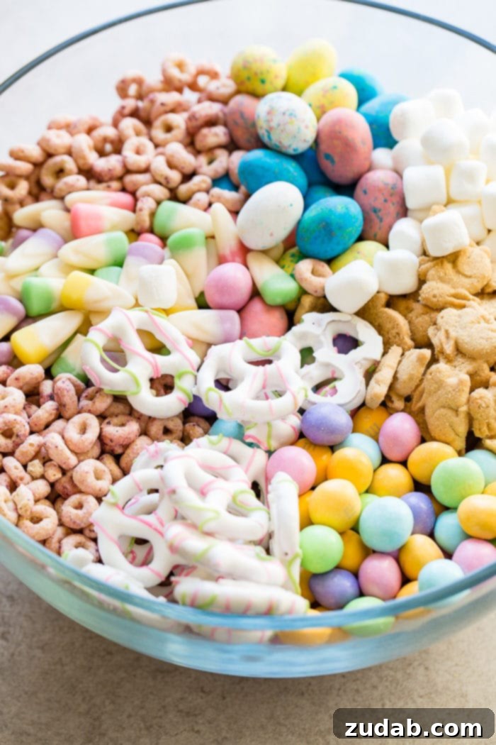 Effortless Easter Snack Medley 6 A close-up of a delightful Easter snack mix in a clear glass bowl, featuring an appealing blend of colorful berry Cheerios, pastel candy corn, chocolate-covered pretzels, speckled malt eggs, Easter M&Ms, and charming bunny cookies.