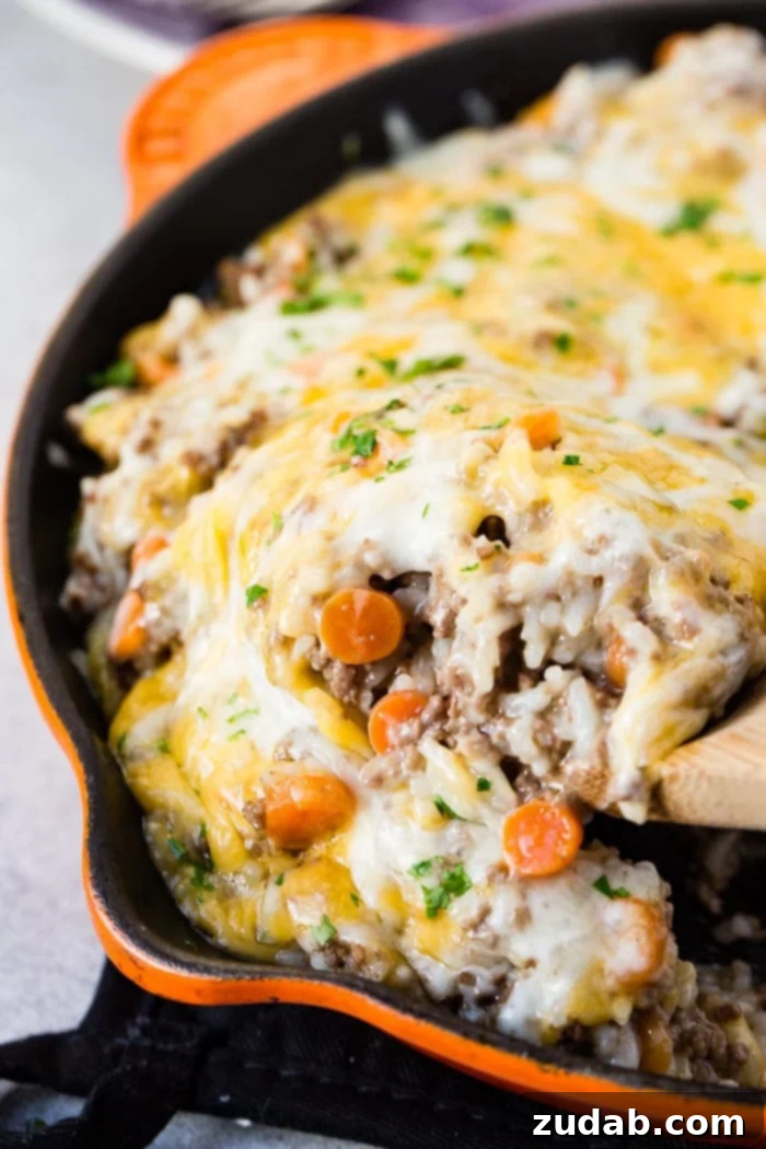 A photo of a scoop of cheesy ground beef and rice casserole being removed from an orange casts iron skillet with a wooden spoon.