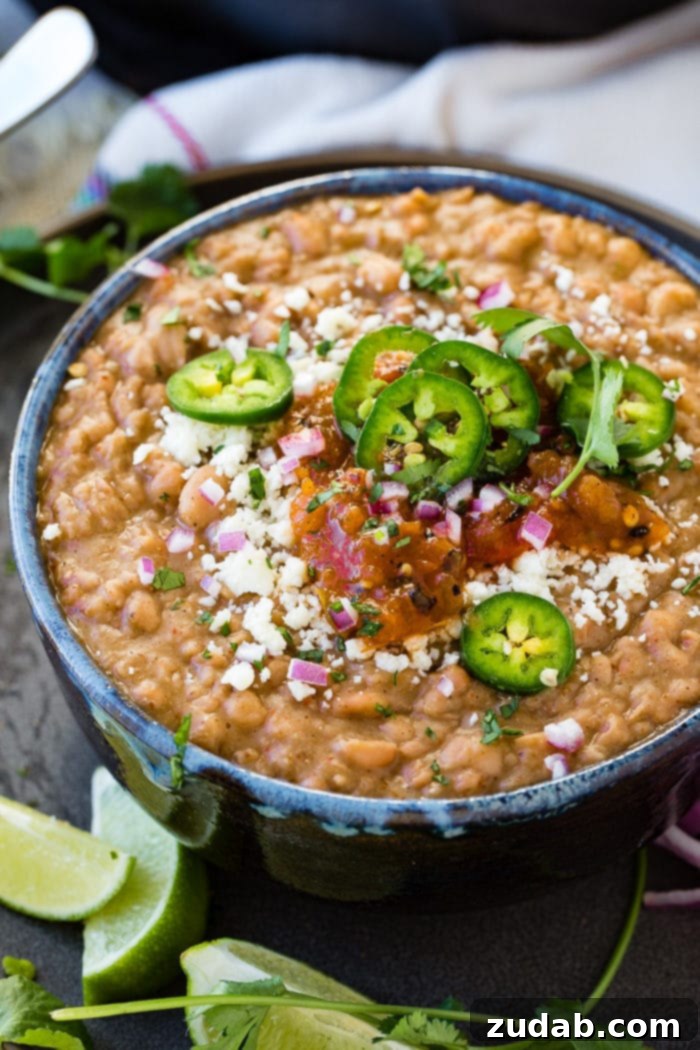Restaurant-Quality Instant Pot Refried Beans 2 A dark blue bowl of creamy Instant Pot refried beans, garnished with fresh sliced jalapeno, diced purple onion, crumbled cotija cheese, and vibrant salsa. A perfect restaurant-style Mexican side dish.
