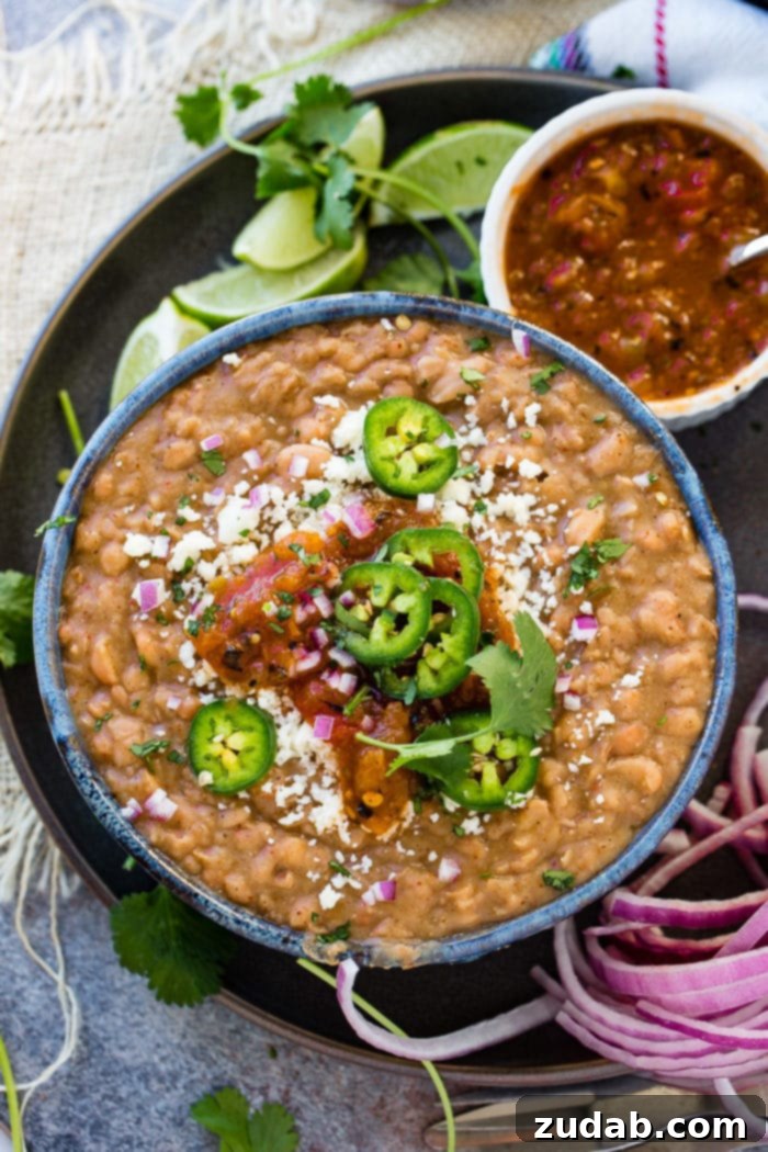 Restaurant-Quality Instant Pot Refried Beans 4 A dark blue bowl of homemade refried beans, generously topped with fresh sliced jalapeno, diced purple onion, crumbled cotija cheese, and a dollop of vibrant salsa. A small cup of salsa sits beside the bowl.