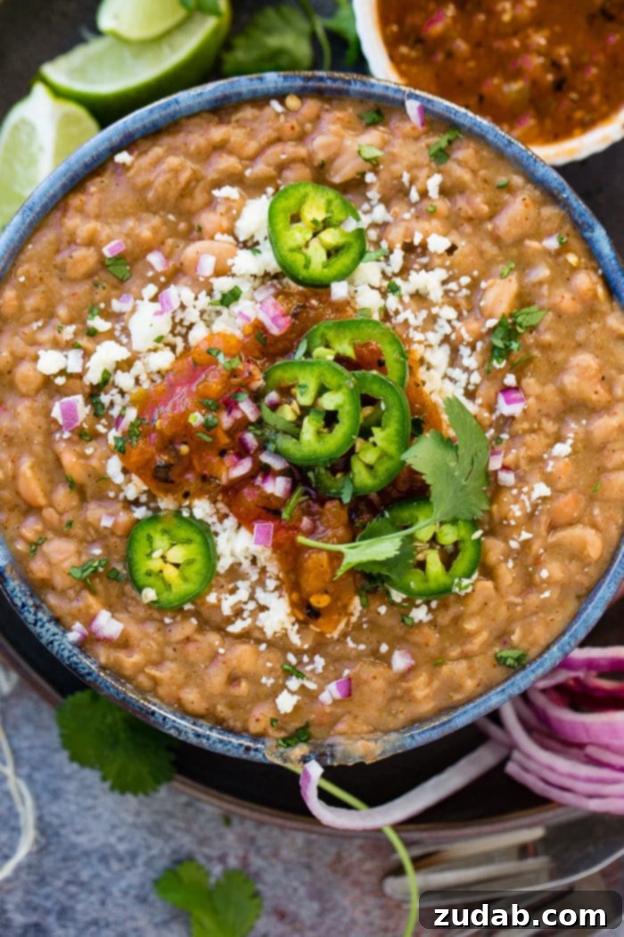 Restaurant-Quality Instant Pot Refried Beans 5 A close-up shot of restaurant-style Instant Pot refried beans, beautifully presented in a dark blue bowl. Garnished with fresh jalapeno slices, diced purple onion, Cotija cheese, and a side of salsa, showcasing their rich texture and vibrant toppings.