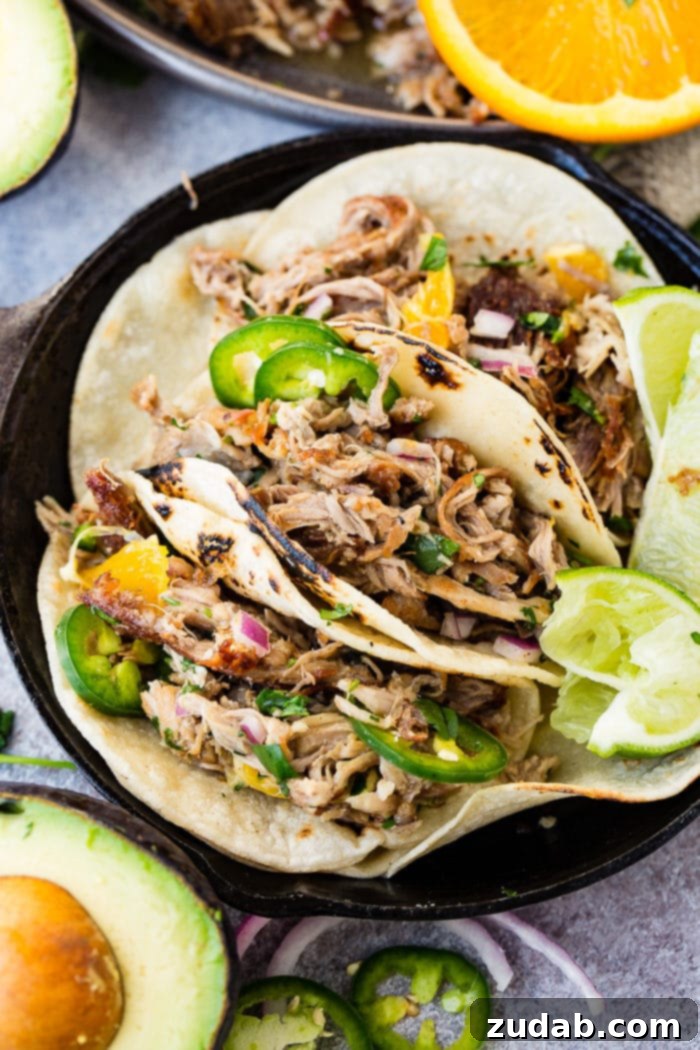 Crispy Tender Pork Carnitas Recipe 2 A photo of three pork carnitas tacos in a cast iron skillet topped with fresh jalapeno slices, diced red onions, and cilantro with a squeezed lime on the side.