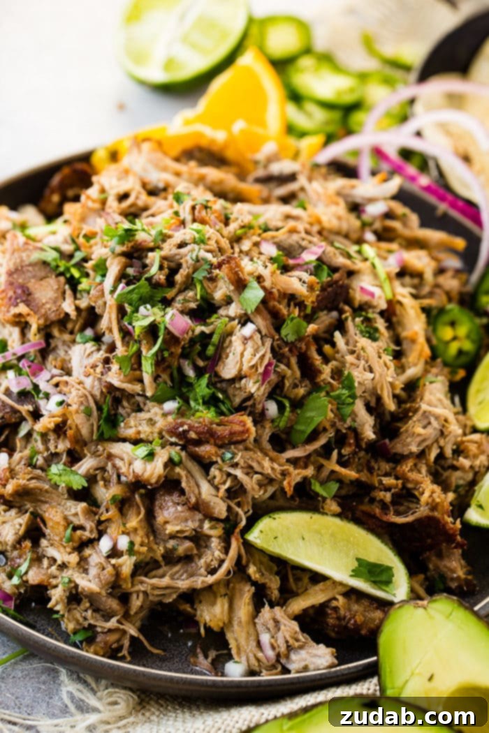 Crispy Tender Pork Carnitas Recipe 3 A photo of a mound of shredded pork carnitas in a cast iron skillet and topped with fresh cilantro, red onions, slices of fresh jalapenos and lime wedges.