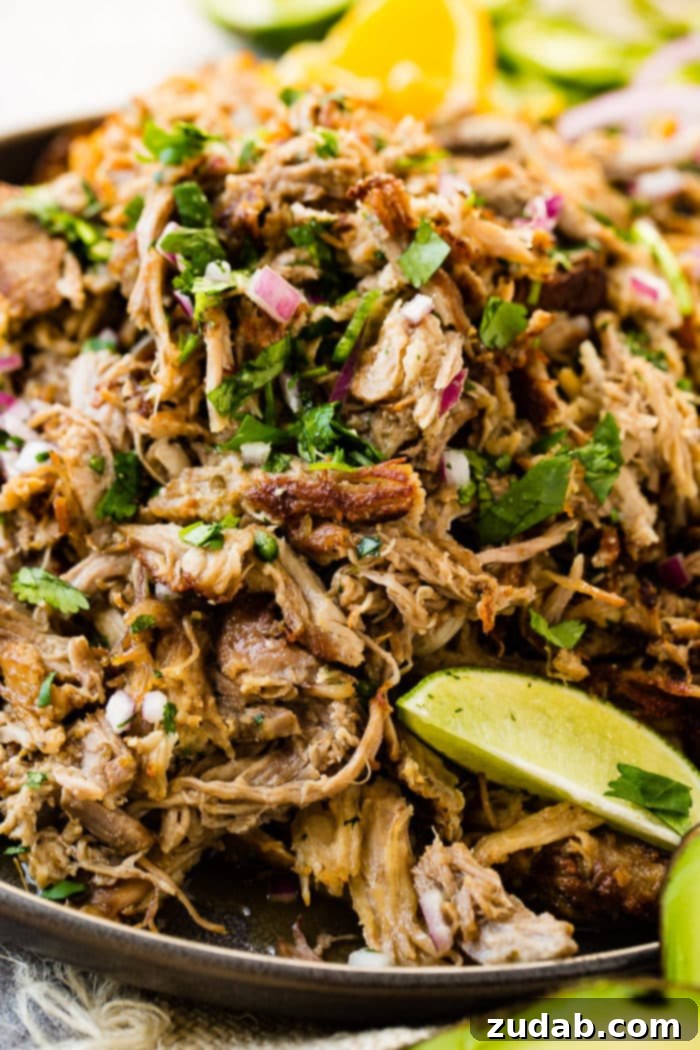 Crispy Tender Pork Carnitas Recipe 4 A photo of a mound of shredded pork carnitas in a cast iron skillet and topped with fresh cilantro, red onions, slices of fresh jalapenos and lime wedges.