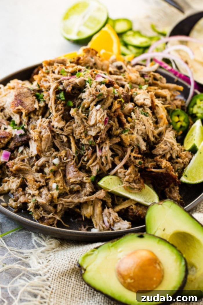 Crispy Tender Pork Carnitas Recipe 6 A photo of a mound of shredded pork carnitas on a dark gray plate and topped with fresh cilantro, red onions, slices of fresh jalapenos and lime wedges.