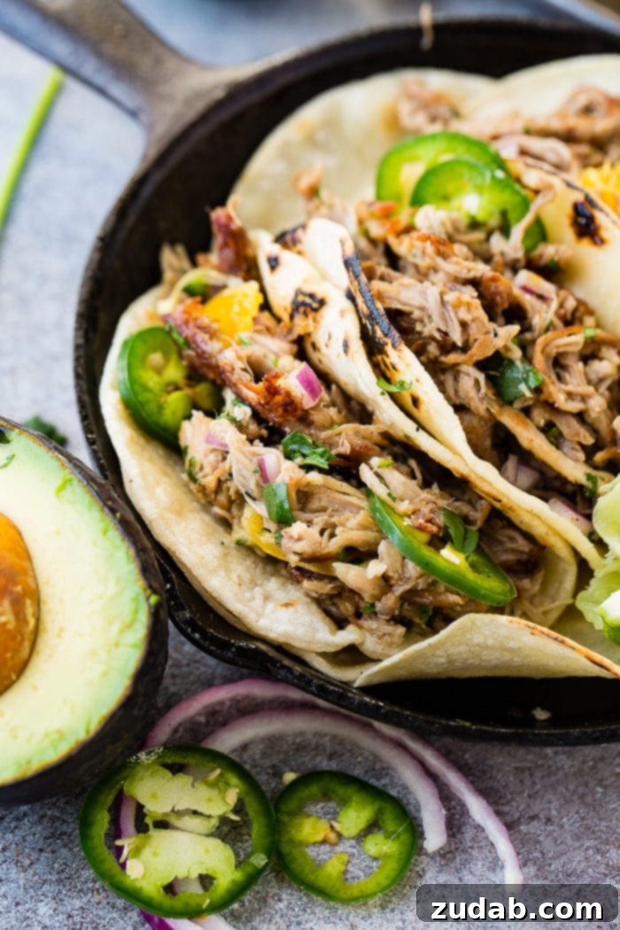 Crispy Tender Pork Carnitas Recipe 7 A photo of three pork carnitas tacos in a cast iron skillet topped with fresh jalapeno slices, diced red onions, and cilantro with a squeezed lime and a halved avocado on the side.