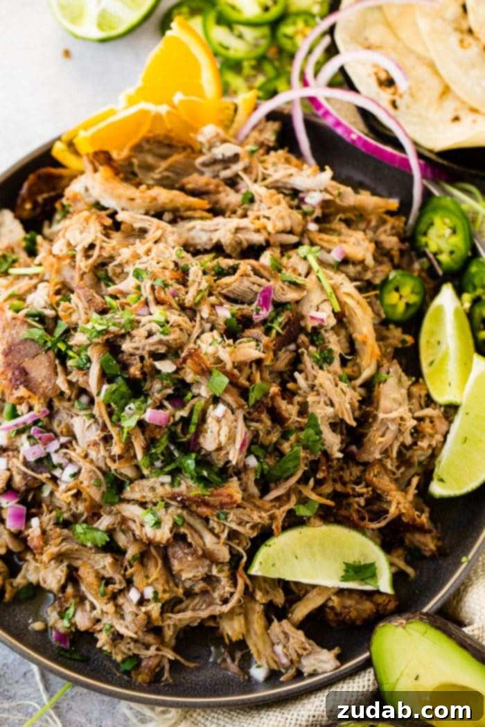 Crispy Tender Pork Carnitas Recipe 8 A photo of a mound of shredded pork carnitas on a dark gray plate and topped with fresh cilantro, red onions, slices of fresh jalapenos and lime wedges.