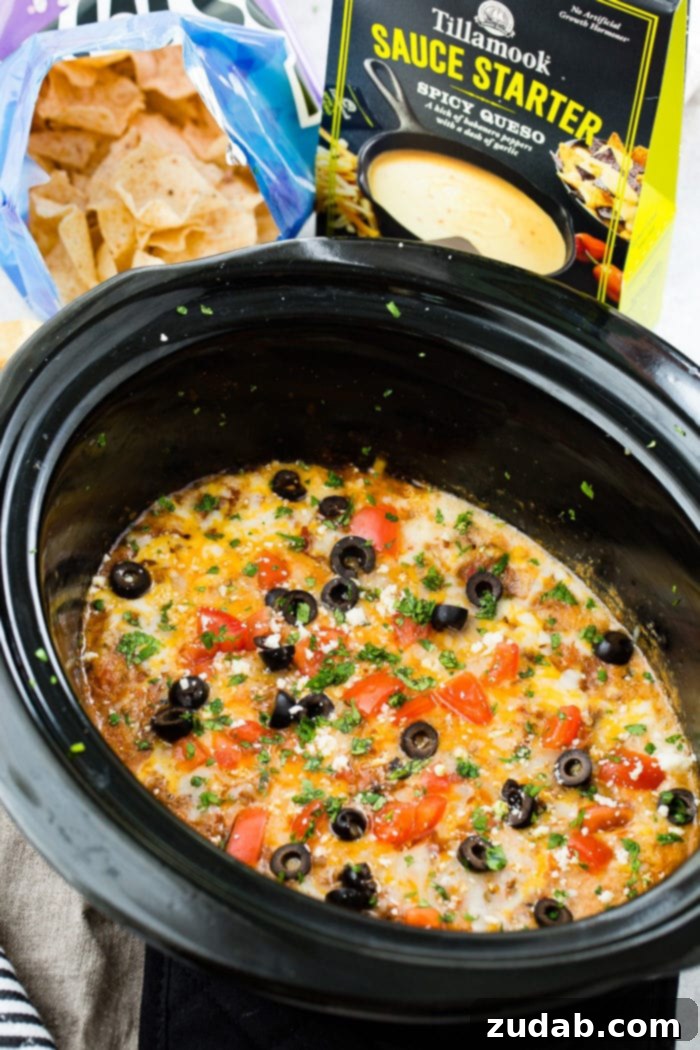 Ultimate Slow Cooker Taco Dip 4 A photo of slow cooker taco dip in a black oval slow cooker topped with melted cheese, sliced olives, chopped tomatoes and cilantro.