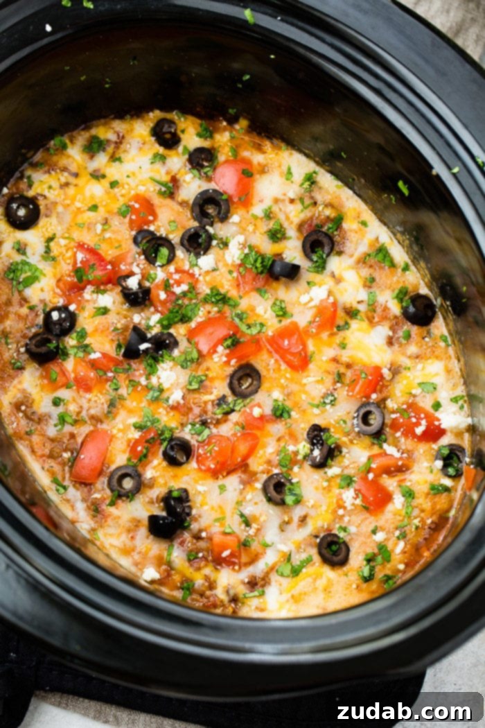 Ultimate Slow Cooker Taco Dip 5 A photo of slow cooker taco dip in a black oval slow cooker topped with melted cheese, sliced olives, chopped tomatoes and cilantro.
