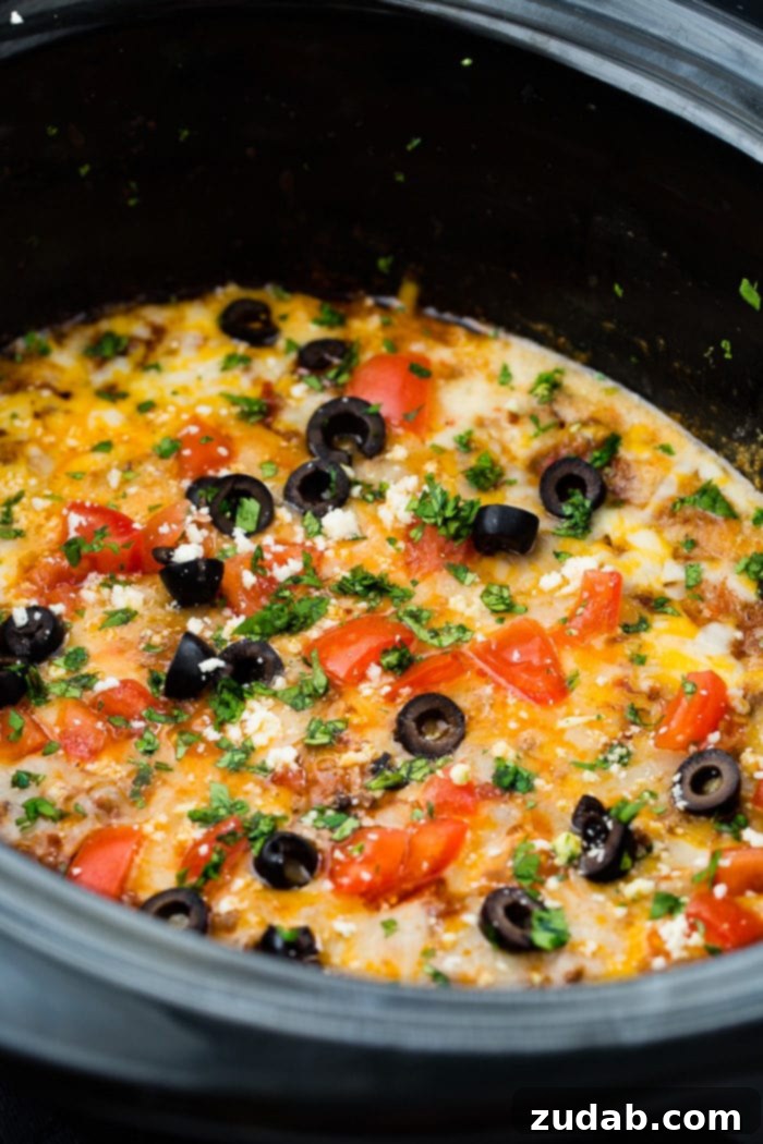 Ultimate Slow Cooker Taco Dip 7 A photo of slow cooker taco dip in a black oval slow cooker topped with melted cheese, sliced olives, chopped tomatoes and cilantro.