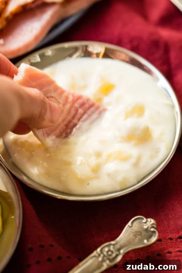 Simple Sauces That Make Ham Shine 7 Two hands elegantly dipping a small, succulent slice of ham into a pristine dish of honey mustard sauce, highlighting the perfect pairing.