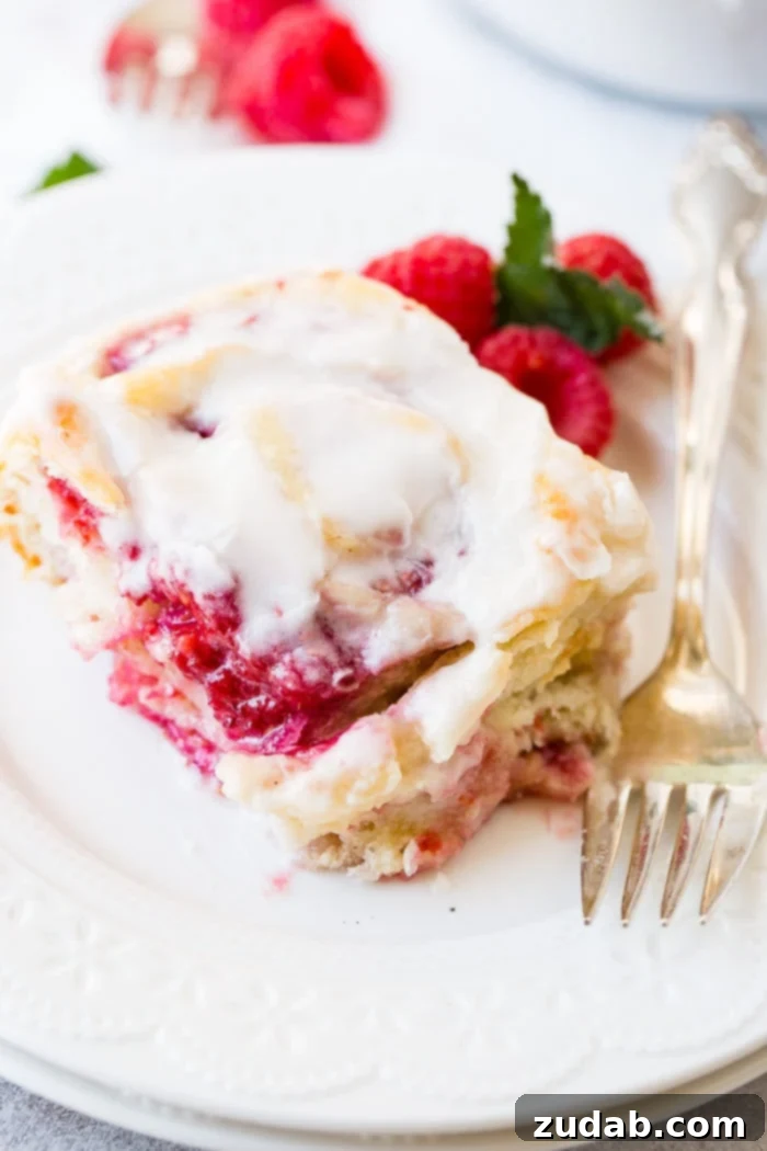 Effortless Raspberry Sweet Rolls 2 A frosted easy raspberry sweet roll on a white plate with a fork laying beside it and fresh raspberries next to it.