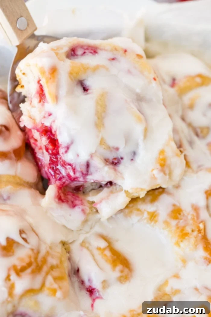 Effortless Raspberry Sweet Rolls 5 A frosted easy berry sweet bread being scooped out of a pan of baked sweet rolls with a spatula.