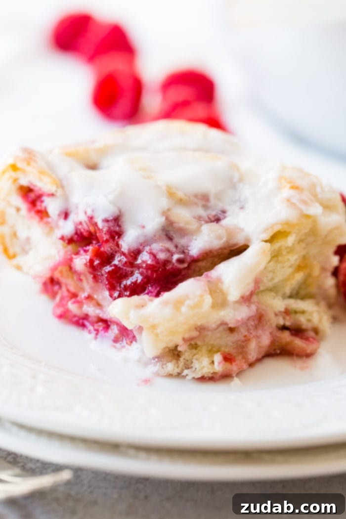 Effortless Raspberry Sweet Rolls 8 A photo of a easy berry sweet swirl roll sitting on a white plate and covered in frosting.