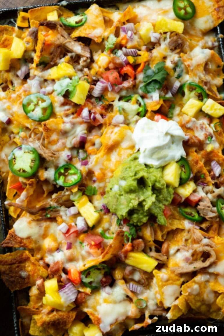 Crockpot Luau Pulled Pork Nachos 6 A vibrant close-up of Hawaiian pulled pork nachos, richly topped with creamy pinto beans, melted cheese, finely diced red onions, fresh chopped cilantro, a dollop of sour cream, and a generous scoop of guacamole.