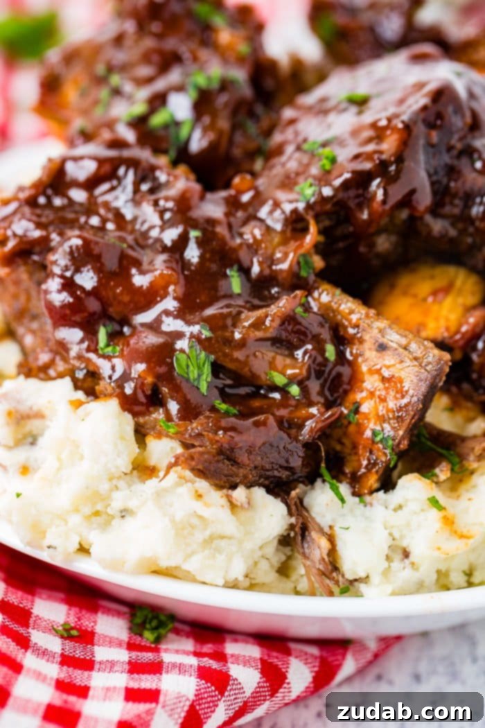 Fall-Off-The-Bone Instant Pot BBQ Short Ribs 3 A photo of sticky bbq beef short ribs piled on top of mashed potatoes on a white plate on top of a red and white checkered cloth napkin.