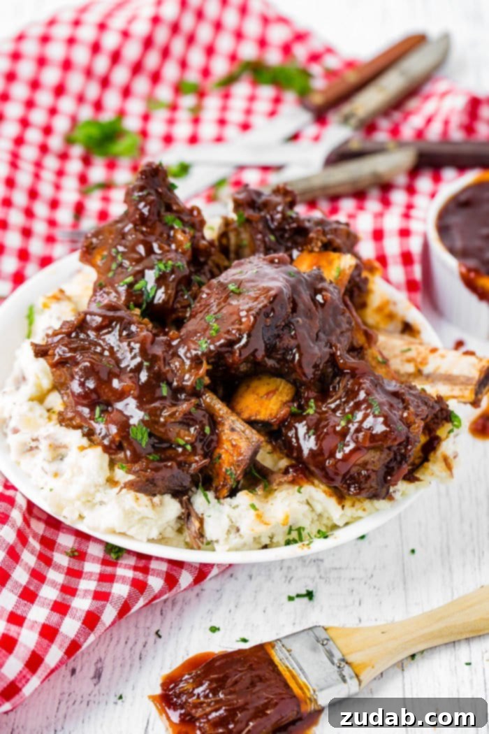 Fall-Off-The-Bone Instant Pot BBQ Short Ribs 4 A photo of sticky bbq beef short ribs piled on top of mashed potatoes on a white plate on top of a red and white checkered cloth napkin.