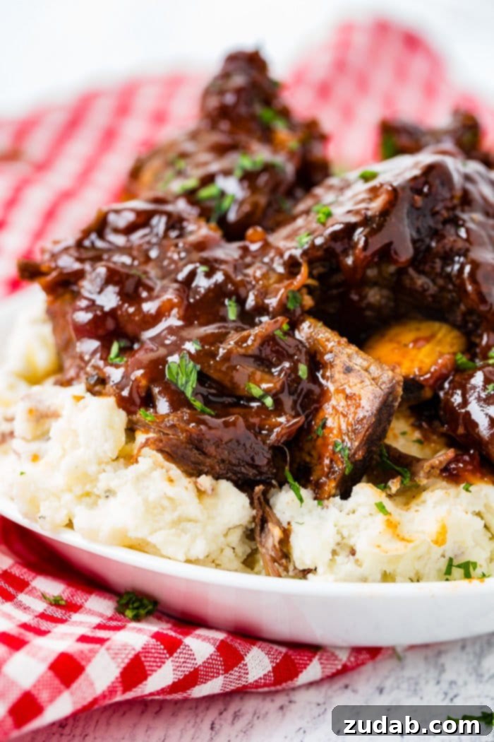 Fall-Off-The-Bone Instant Pot BBQ Short Ribs 5 A photo of sticky bbq beef short ribs piled on top of mashed potatoes on a white plate on top of a red and white checkered cloth napkin.