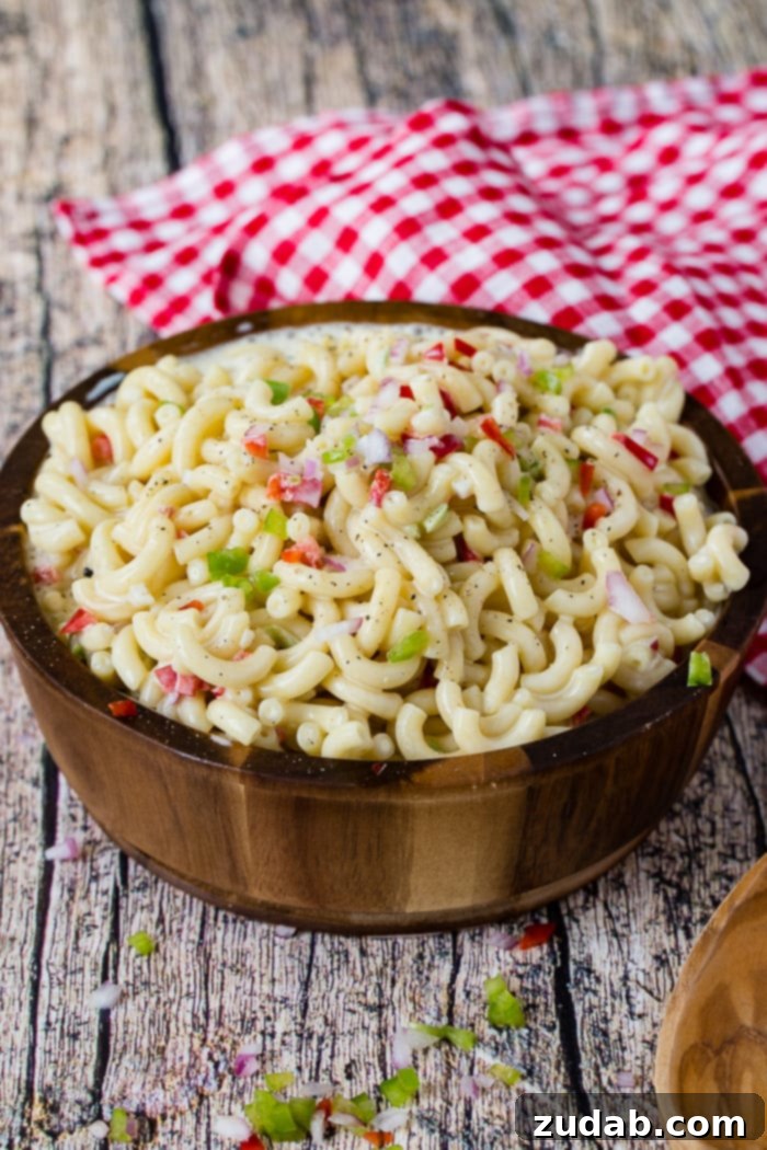 The Ultimate Macaroni Pasta Salad Recipe 4 A wooden bowl on a red and white checkered napkin full of creamy cold noodles and vegetables.