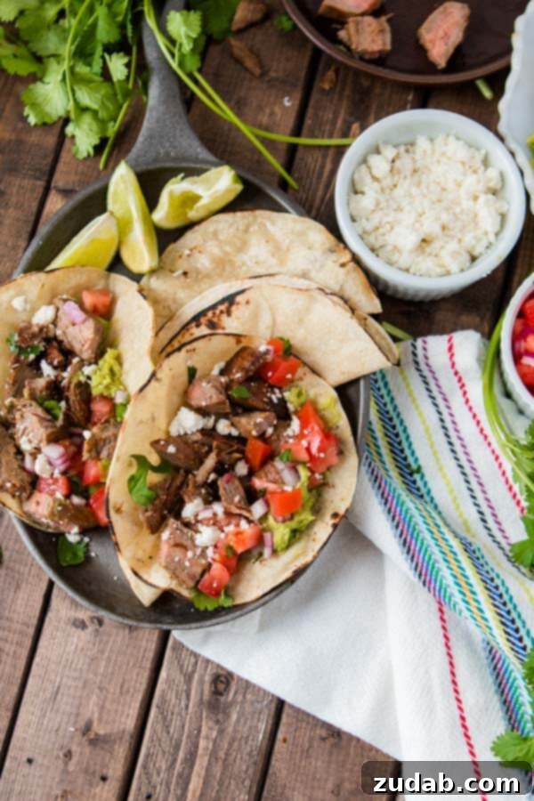 A plate of two delicious carne asada tacos with two tortillas and three slices of lime with a small white bowl of cheese on the side.