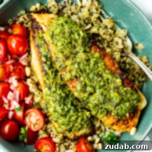 a photo of two golden tilapia filets topped with bright pesto sauce on a bed of quinoa with grape tomatoes on the side