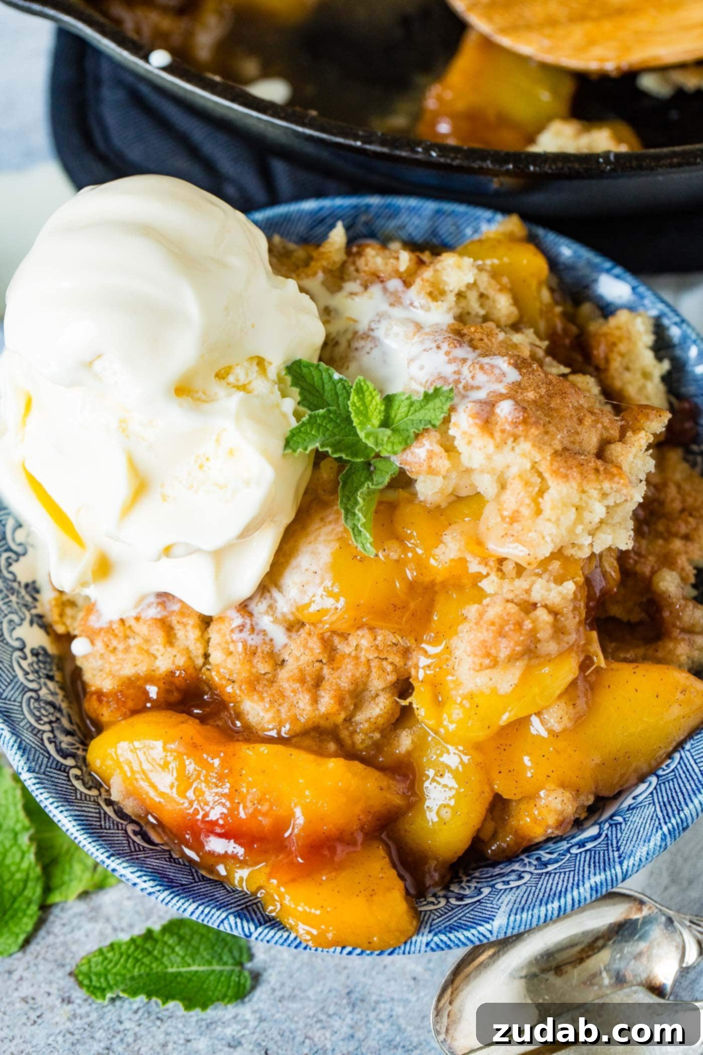 A photo of a bowl of fresh peach cobbler with a scoop of vanilla ice cream on top and a sprig of fresh mint.