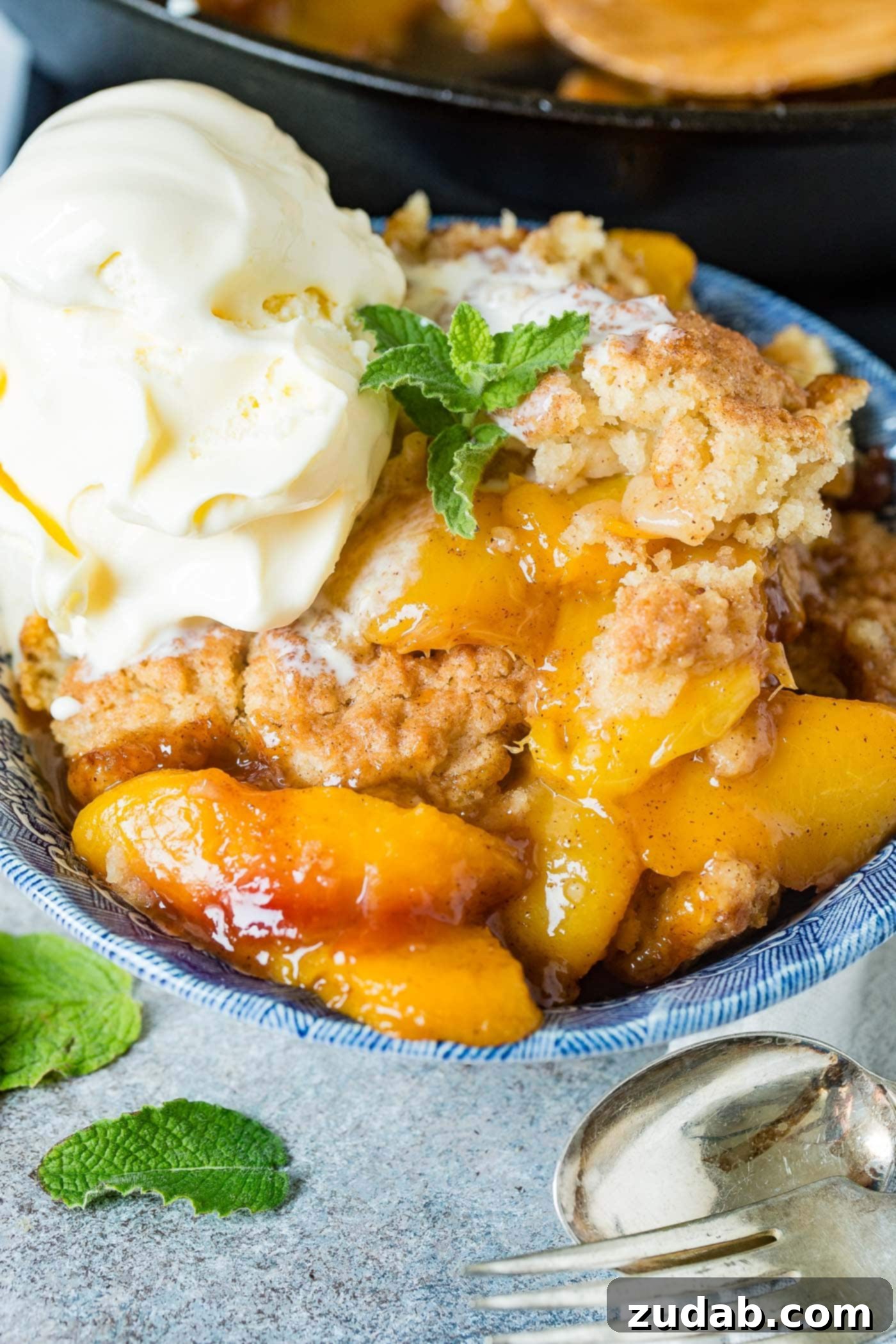 A photo of a bowl of fresh peach cobbler with a scoop of vanilla ice cream on top and a sprig of fresh mint.