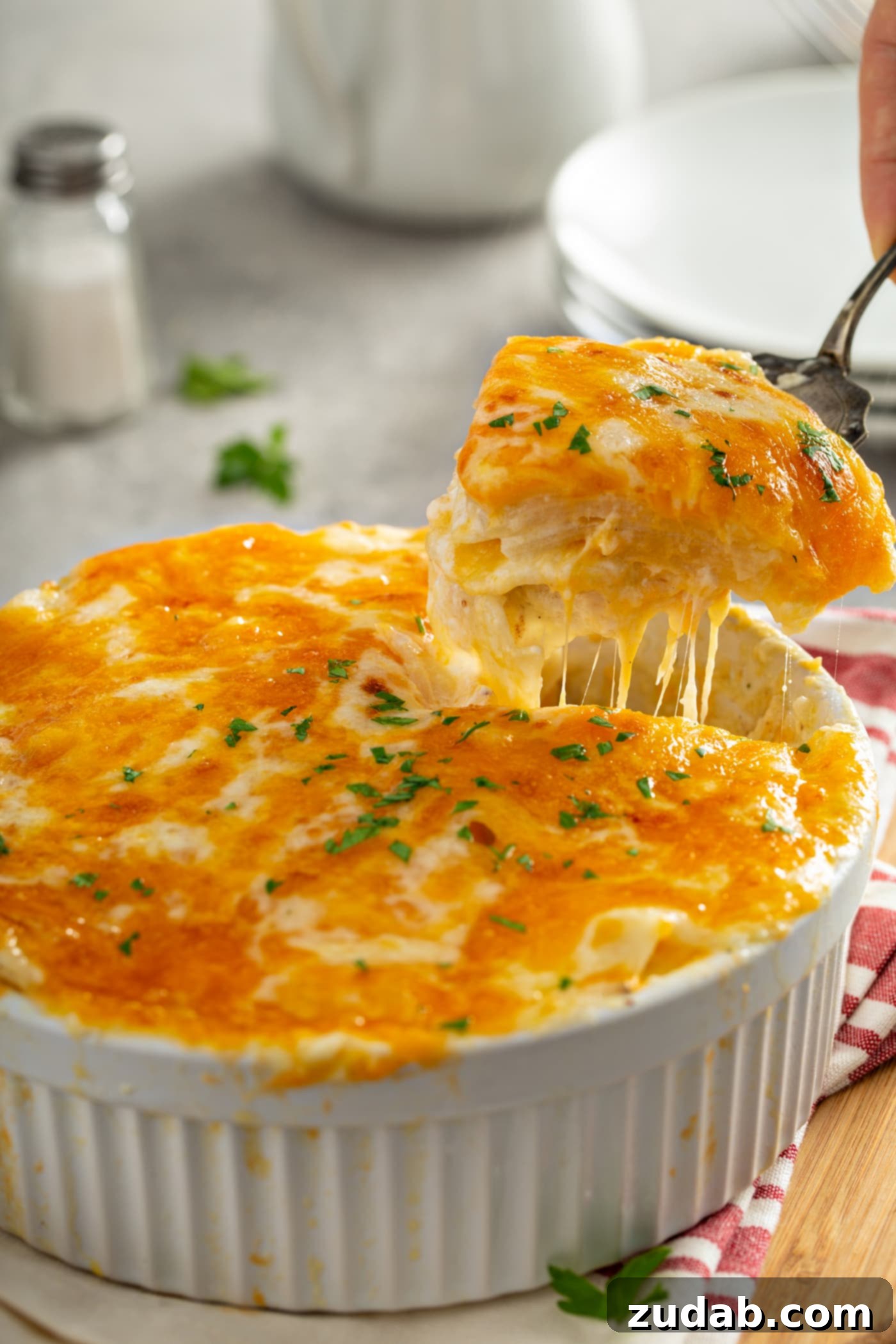 Simple Instant Pot Cheesy Potatoes A serving of instant pot cheesy potatoes are being lifted from a white casserole dish with a serving spoon. the melted cheese is stringing out below the spoon. there is a stack of dinner plates in the background and a red and white kitchen towel beside the casserole dish.