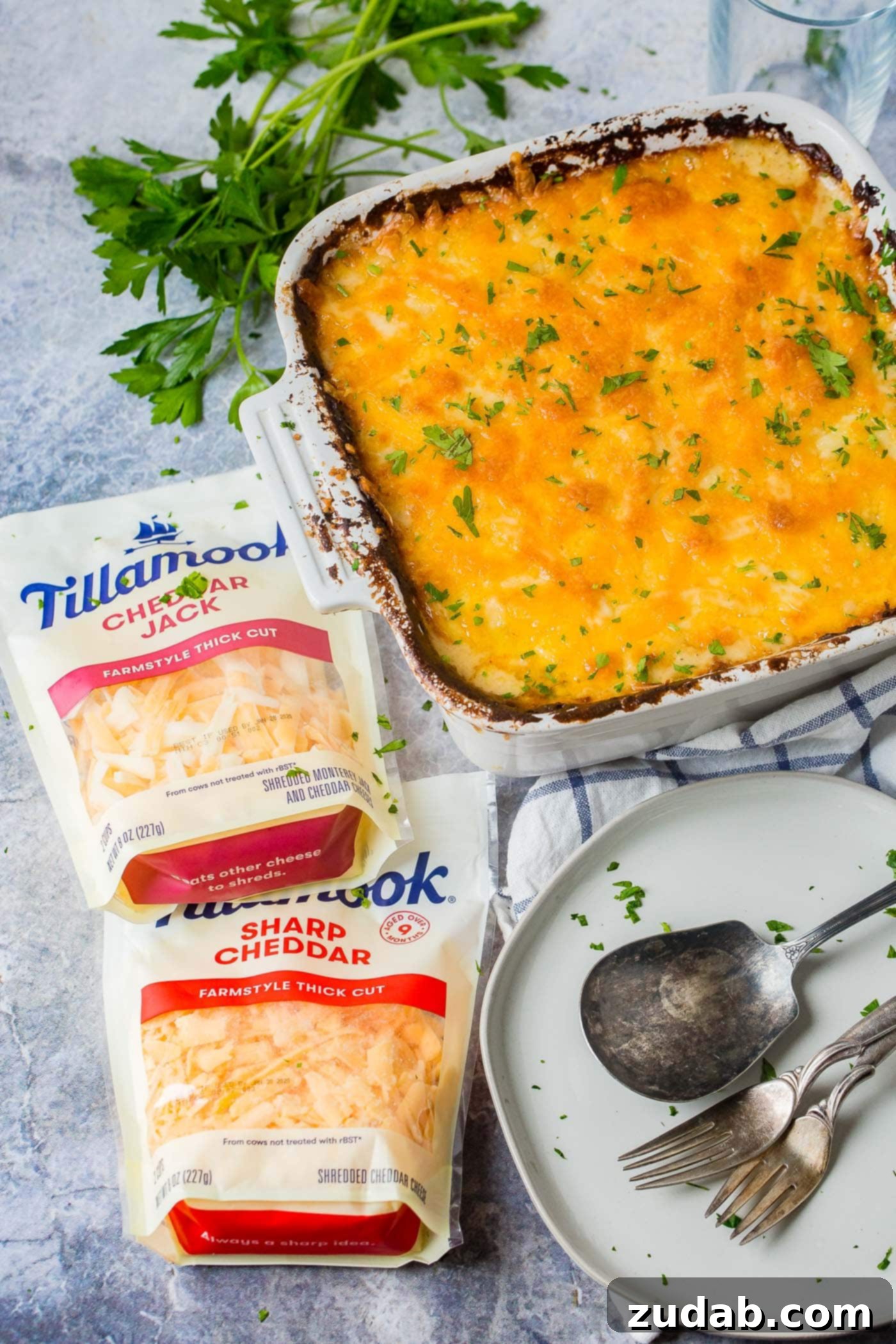 The Best Cheesy Potato Casserole Side Dish 4 baking dish of cheesy potato casserole next to two bags of shredded cheese and a grey plate with utensils