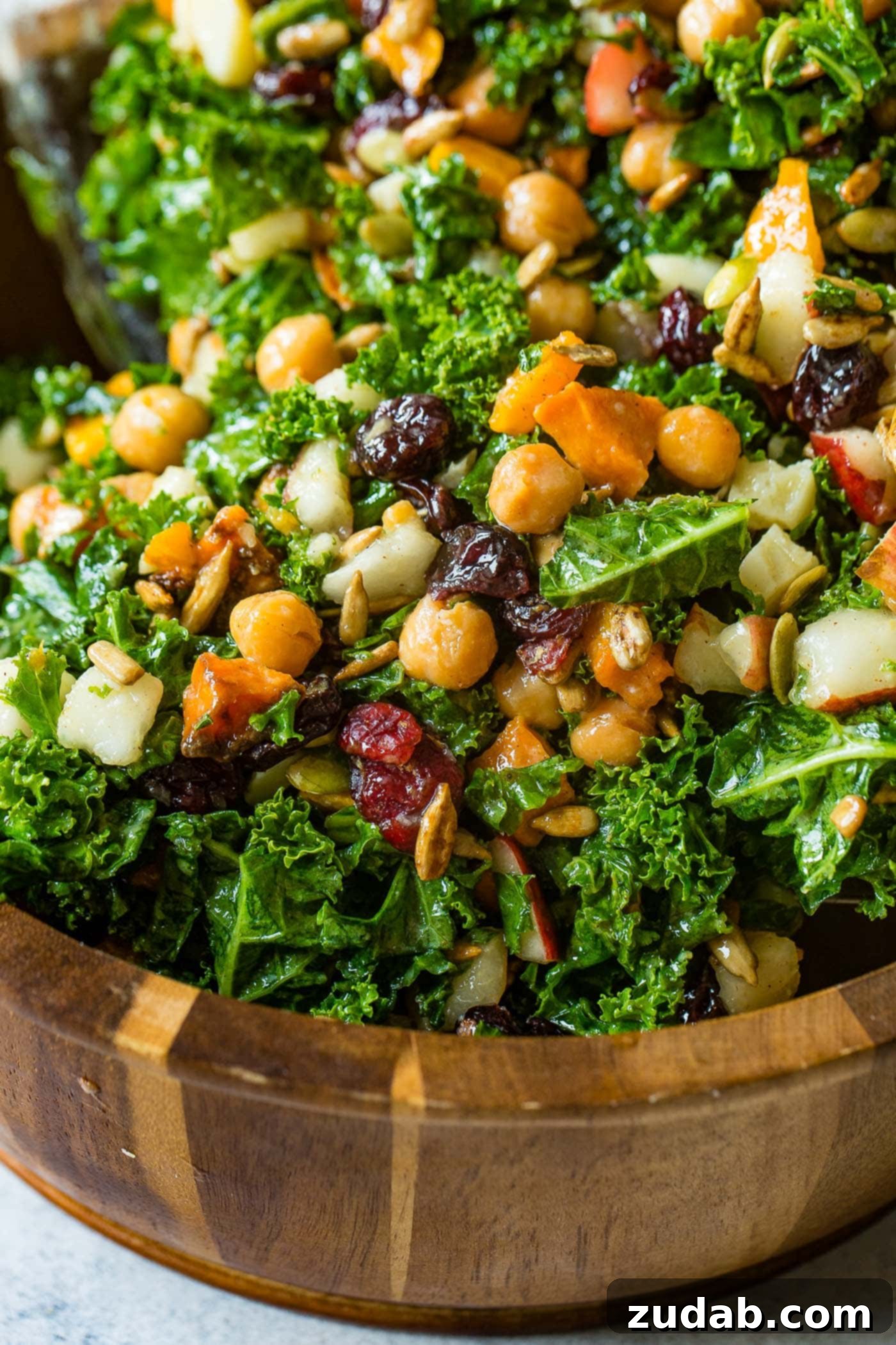 Winter Roasted Butternut Squash Kale Salad A photo of a large wooden bowl full of kale salad topped with roasted butternut squash, craisins, chickpeas, pepitas, and gouda cheese.
