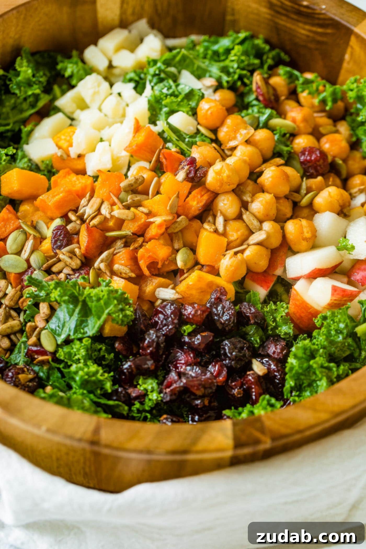 Winter Roasted Butternut Squash Kale Salad A photo of a large wooden bowl full of kale salad topped with roasted butternut squash, craisins, chickpeas, pepitas, and gouda cheese.