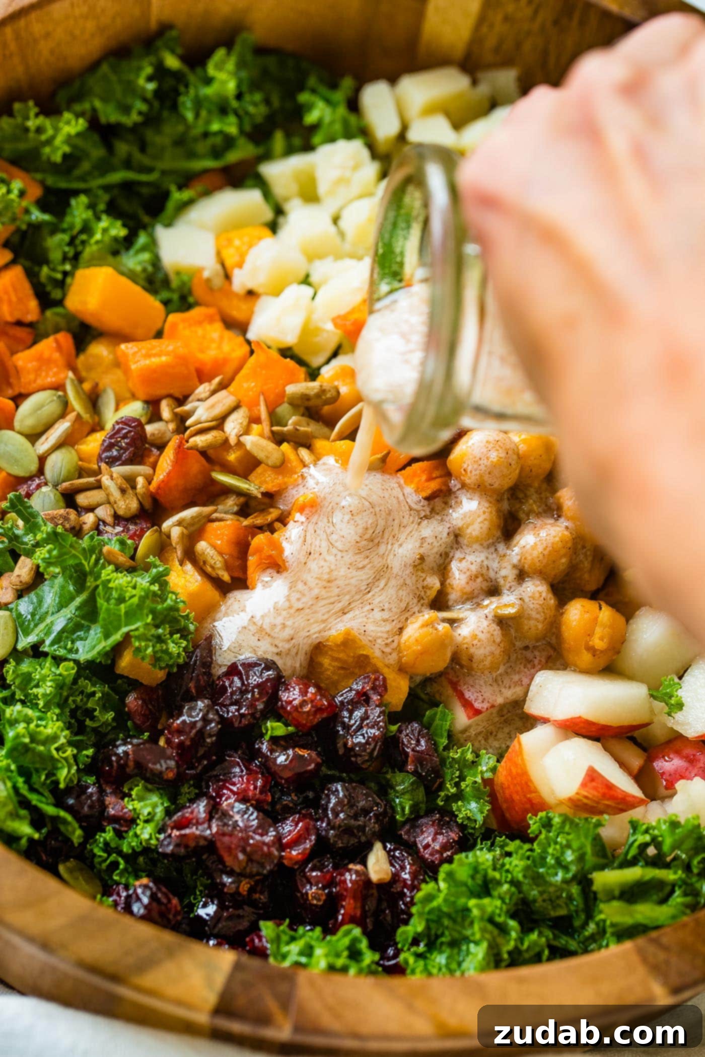 Winter Roasted Butternut Squash Kale Salad A photo of dressing being poured on a large wooden bowl full of kale salad topped with roasted butternut squash, craisins, chickpeas, pepitas, and gouda cheese.