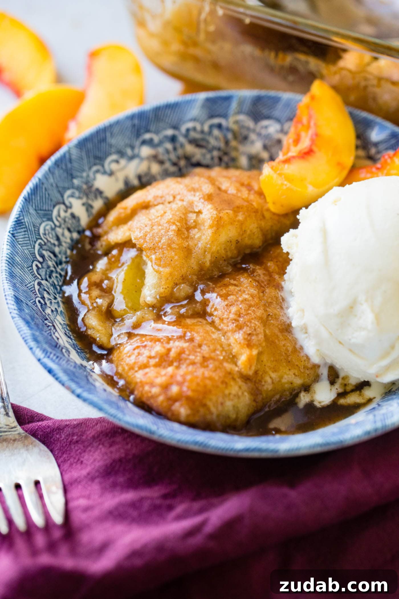 How to Make Peach Dumplings with Crescent Rolls and Sprite a blue dish on a purple napkin with two peach dumplings and a scoop of creamy vanilla ice cream