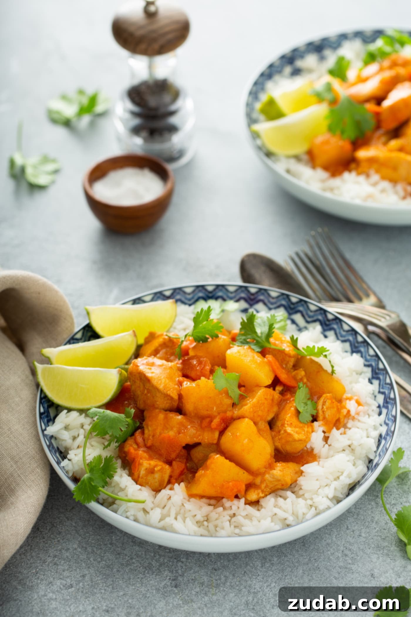 Chicken Curry Recipe in the Instant Pot A bowl of white rice with chicken curry on top. There are chunks of chicken, potatoes and carrots in a curry sauce. Three lime wedges are on the edge of the bowl and fresh parsley is sprinkled on top. There are forks and spoons next to the bowl. In the background is a container of salt and pepper and another serving of chicken curry.