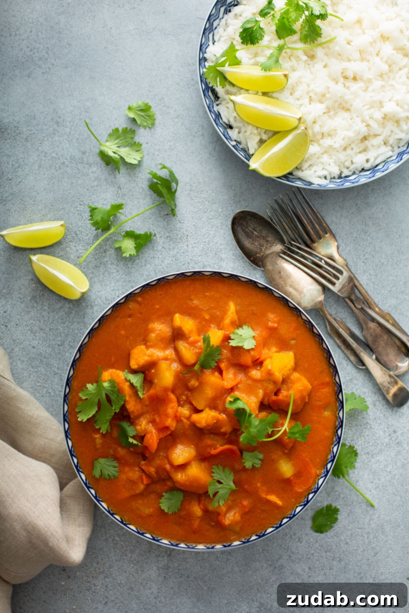 Chicken Curry Recipe in the Instant Pot A bowl of instant pot chicken curry with chunks of chicken, carrots and potatoes. Fresh parsley leaves are scattered on top and on the table. there are lime wedges and silver ware in the background.