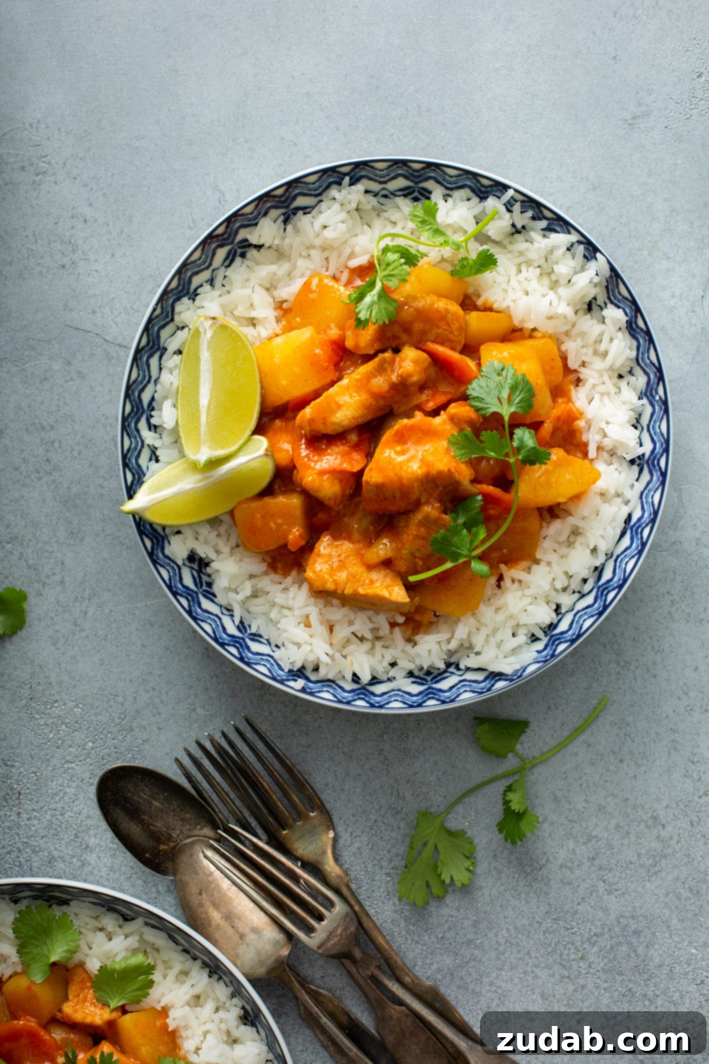 Chicken Curry Recipe in the Instant Pot A bowl of rice with chicken curry on top. There are fresh parsley leaves sprinkled on top and two lime wedges on the edge of the rice. Spoons and forks are on the table next to the bowl.