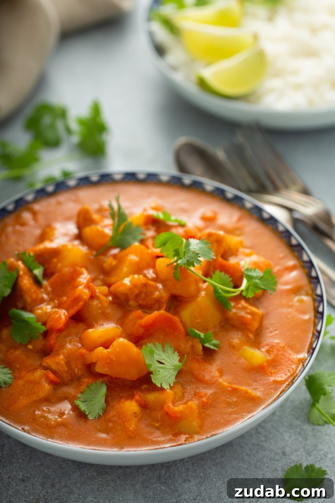 Chicken Curry Recipe in the Instant Pot A bowl of chicken curry with chunks of chicken and potatoes and carrots in a curry sauce. Parsley leaves are scattered on top and on the table next to the curry. A fork and spoon and a plate with lime wedges are in the background.