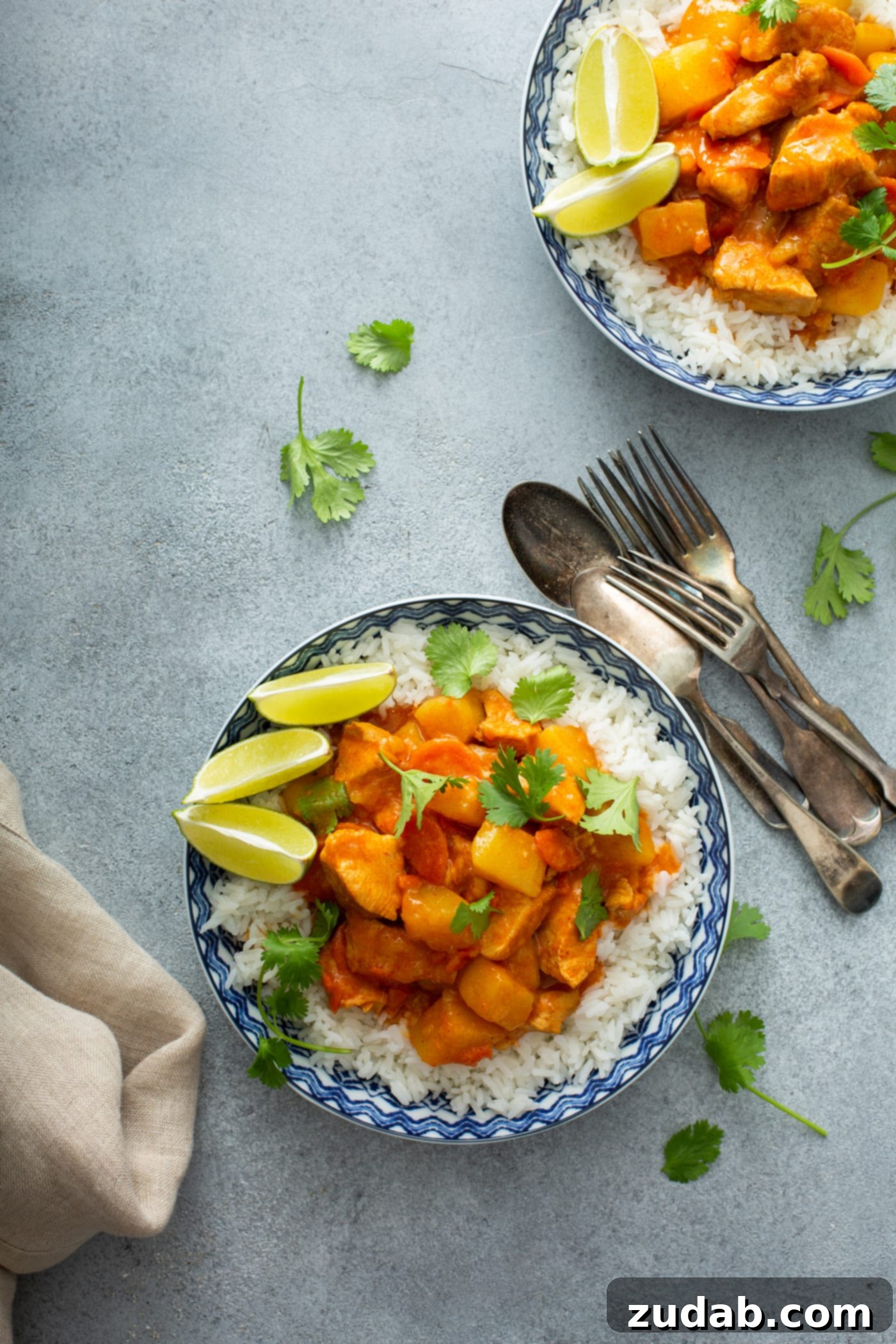 Chicken Curry Recipe in the Instant Pot A bowl of rice topped with chicken curry. There are lime wedges on the edge of the bowl and forks and spoons next to the bowl. Another bowl of curry is in the background.