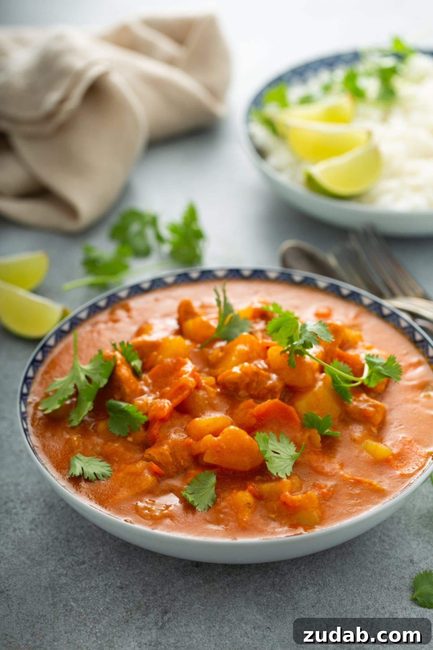 Chicken Curry Recipe in the Instant Pot A large bowl of chicken curry. There are chunks of chicken and potatoes in a bright curry sauce. Parsley leaves and lime wedges are in the background, and a fork and spoon are next to the bowl of curry.