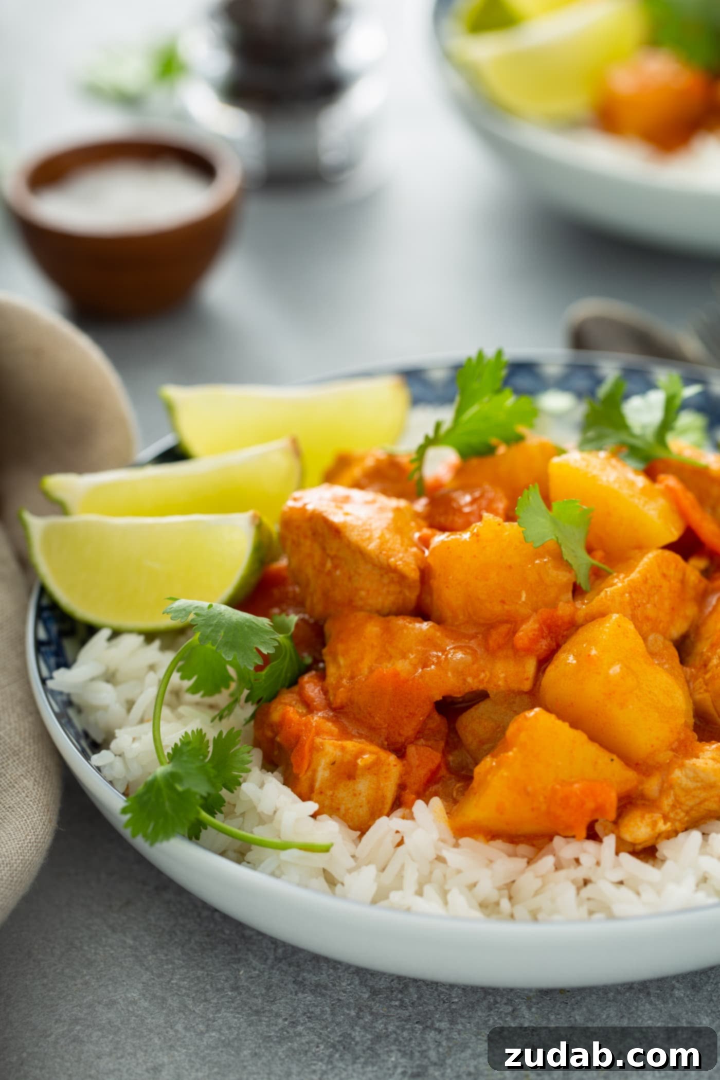 Chicken Curry Recipe in the Instant Pot A bowl of white rice topped with chicken curry. There are chunks of chicken, potatoes and carrots in a curry sauce. It is sprinkled with fresh parsley and there are three lime wedges on the edge of the bowl. A container of salt and pepper are in the background.