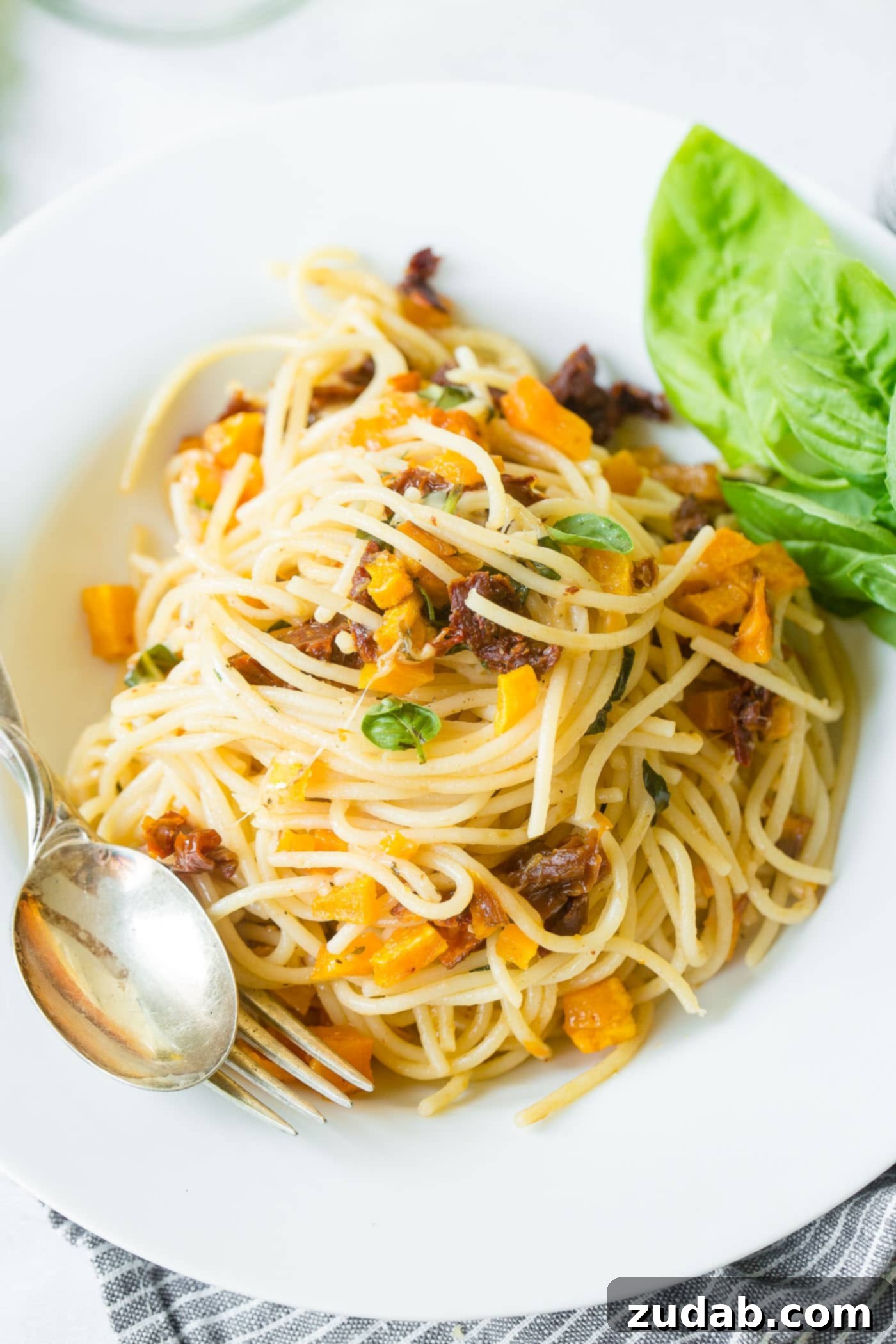 25 Minute Vegetarian Butternut Squash Pasta A photo of a serving of butternut Squash Pasta in a white bowl with a fork and spoon sitting on the side of the bowl. The pasta is vibrant with orange squash, red tomatoes, and green basil, hinting at a delicious, easy dinner.