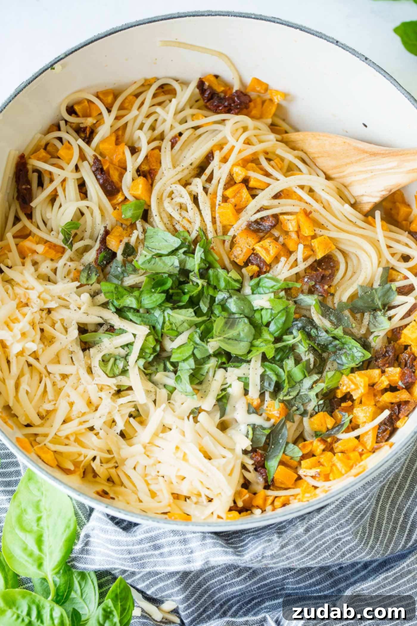 25 Minute Vegetarian Butternut Squash Pasta A photo of a large pot filled with cooked spaghetti, generously mixed with chopped fresh basil, golden roasted butternut squash cubes, melted cheese, and vibrant sun-dried tomatoes, ready to be served.